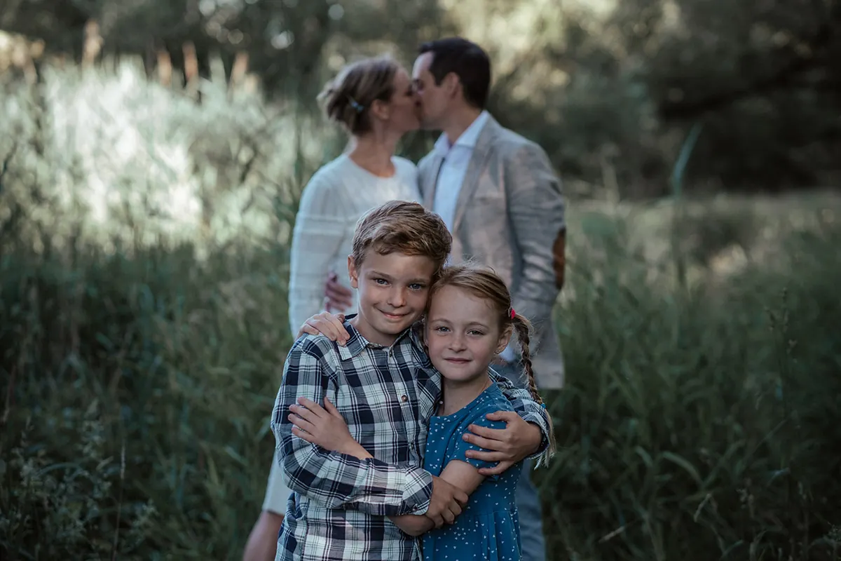 Bruder und Schwester umarmen sich lachend, im Hintergrund küssen sich die Eltern in hohem Gras