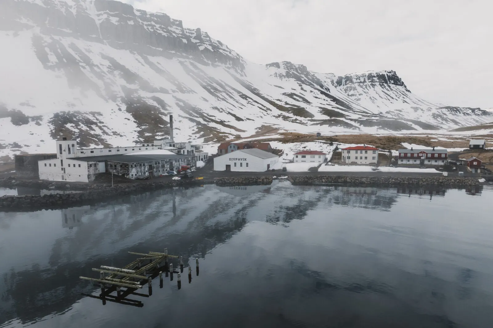 Die alte Heringsfabrik von Djúpavík spiegelt sich im Fjord, dahinter verschneite Berge
